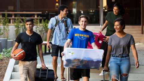 Group of five people moving into Grafton Hall. They are carrying suitcases and boxes, chatting and smiling.